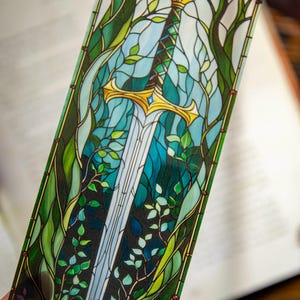 Peut inclure: Un marque-page en verre coloré représentant une épée avec une poignée dorée, sur un fond bleu et vert. L'épée est entourée de feuilles et de vignes vertes.