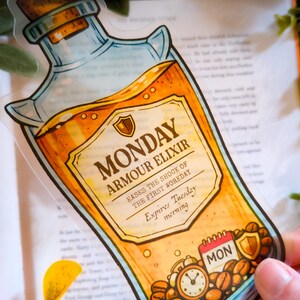 Puede incluir: Adhesivo transparente con una ilustración de dibujos animados de una botella de poción con la etiqueta "Monday Armour Elixir". La botella contiene un líquido dorado y tiene un tapón de corcho. La etiqueta incluye el texto "Alivia el impacto del primer día de trabajo".