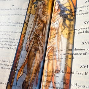 May include: A tall, narrow bookmark featuring a translucent illustration of a figure with long dark hair, wearing layered tan and orange garments. The design incorporates arched architectural lines and warm amber tones, placed between the pages of a book.