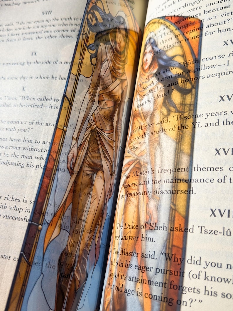 May include: A tall, narrow bookmark featuring a translucent illustration of a figure with long dark hair, wearing layered tan and orange garments. The design incorporates arched architectural lines and warm amber tones, placed between the pages of a book.