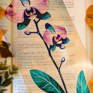 May include: A transparent bookmark featuring a detailed illustration of an orchid plant with two blooming pink and purple flowers, several small buds, and dark green leaves with visible veins, set against a clear, smooth rectangular surface.