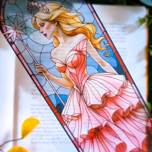 Peut inclure: Un marque-page rectangulaire présentant une illustration de style vitrail d'une silhouette blonde portant une robe rose à volants et une couronne. La silhouette tient un sceptre étoilé sur un fond bleu avec des lignes de plomb noires.