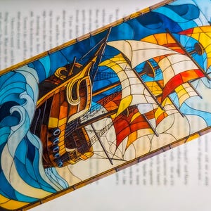Op de afbeelding: Een kleurrijke boekenlegger met een zeilschip met wapperende zeilen. Het kunstwerk toont het schip op een golvende zee, met een blauwe lucht en gele, oranje en witte zeilen. De boekenlegger ligt op een open boek.