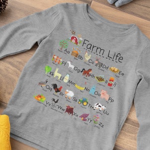 May include: A gray long-sleeved shirt with a farm-themed alphabet design. The shirt features a red barn, animals, and the letters A through Z. The text "Farm Life" is printed on the shirt.