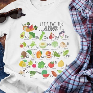 May include: White t-shirt with a graphic of a colorful alphabet made of fruits and vegetables. The text "LET'S EAT THE ALPHABET!" is printed above the graphic.