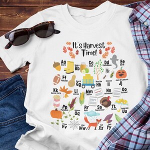 May include: White t-shirt with a colourful alphabet chart featuring autumn-themed illustrations. The chart includes the text "It's Harvest Time!" and depicts various autumnal items like pumpkins, acorns, leaves, and rain clouds.