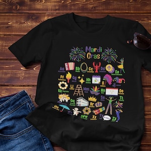 May include: Black t-shirt with a Mardi Gras alphabet design. The letters are illustrated with images of Mardi Gras traditions, such as beads, masks, and king cakes. The text "Mardi Gras" is at the top of the design.
