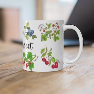 Könnte beinhalten: Weiße Keramiktasse mit der Aufschrift 'Sweet' in Schwarz und Illustrationen verschiedener Beeren, darunter Erdbeeren, Blaubeeren, Kirschen und Johannisbeeren.
