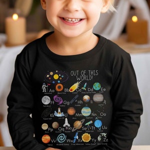 May include: A black long-sleeved shirt with a space-themed alphabet design. Each letter is paired with a space-related image, such as a planet, star, or astronaut. The shirt has the text "OUT OF THIS WORLD!" printed on it.