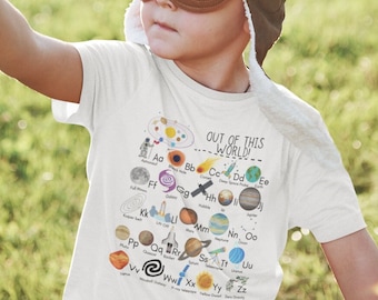 Könnte beinhalten: Ein kleines Kind trägt eine braune Fliegermütze und eine Schutzbrille und hält ein Spielzeugflugzeug in der Hand. Das Kind trägt ein weißes T-Shirt mit einer Grafik von Planeten und dem Text "OUT OF THIS WORLD".