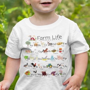 May include: A young child wearing a white t-shirt with a farm-themed alphabet design. The shirt features illustrations of farm animals and objects, such as a cow, pig, chicken, tractor, and barn. The text "Farm Life" is printed at the top of the shirt.
