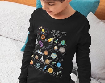 Könnte beinhalten: Ein Kind trägt ein schwarzes Langarmshirt mit einem Planeten-Motiv und dem Text "OUT OF THIS WORLD!" Das Shirt zeigt eine bunte Illustration von Planeten und Sternen mit Buchstaben des Alphabets. Das Kind sitzt auf einem Bett und schaut in ein Buch.