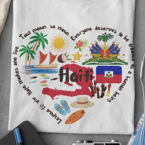 May include: White t-shirt with a graphic design featuring a map of Haiti, the Haitian flag, and the text "Tout moun. se moun. Everyone deserves to be treated as a human being. Lespwa f&egrave; viv. Hope makes one live."