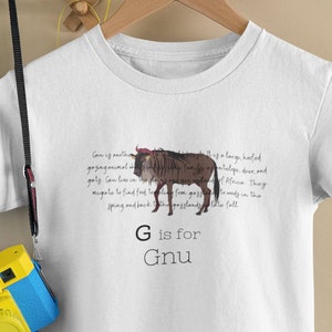 Puede incluir: Camiseta blanca con una ilustración marrón y blanca de un GNU, un gran animal de pezuña que pasta en las llanuras de África. El texto "G is for GNU" está impreso debajo de la ilustración.