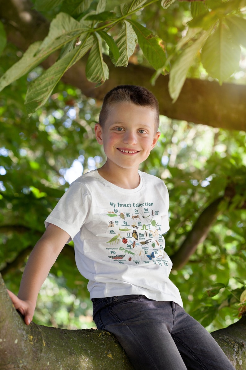 Op de afbeelding: Een jonge jongen draagt een wit T-shirt met een kleurrijke insectenillustratie en de tekst "My Insect Collection". Hij zit op een boomtak buiten.