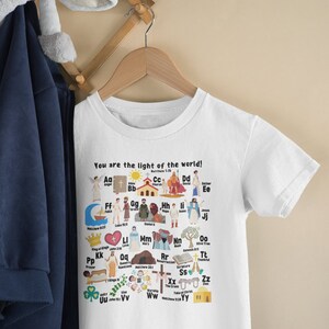 May include: White t-shirt with a colorful alphabet design featuring illustrations of Christian themes. Each letter is paired with a word and a Bible verse. The text at the top reads "You are the light of the world!"