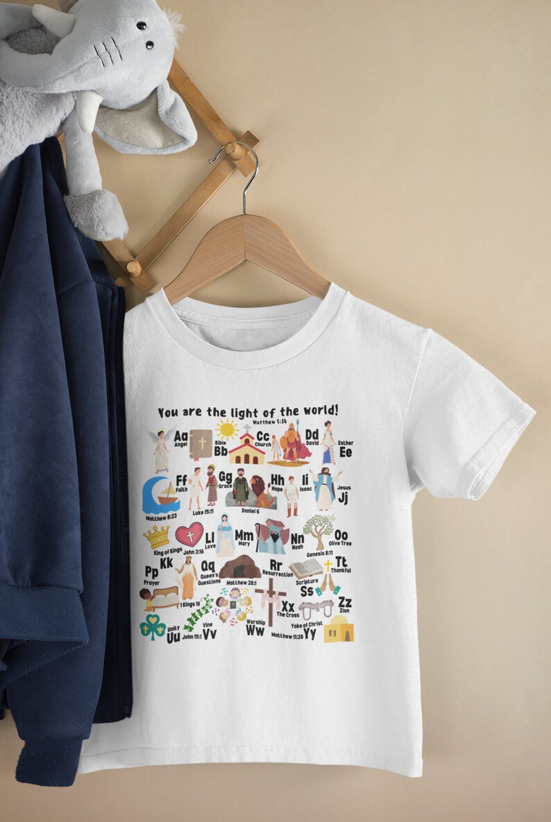 May include: White t-shirt with a colorful alphabet design featuring illustrations of Christian themes. Each letter is paired with a word and a Bible verse. The text at the top reads "You are the light of the world!"