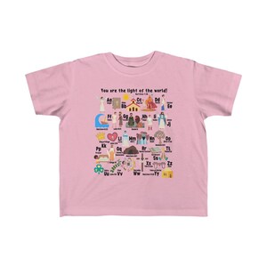 May include: A pink children's t-shirt with a colorful alphabet design featuring illustrations of religious figures and symbols. The text "You are the light of the world!" is printed at the top of the shirt.  Matthew 5:14