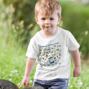 May include: A young child wearing a white t-shirt with the text "My Insect Collection" and various insect illustrations. The child is standing in a grassy area, wearing blue jeans.