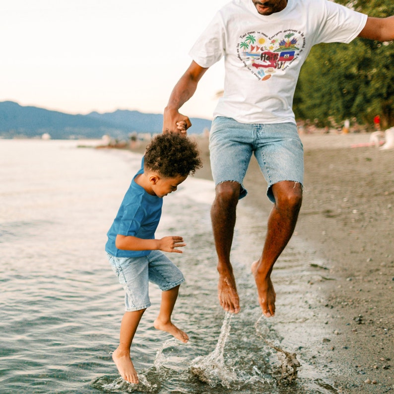 May include: A father and son are jumping in the water at the beach. The father is wearing a white t-shirt with a colorful heart design and blue denim shorts. The son is wearing a blue t-shirt and blue denim shorts.