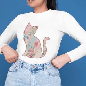 May include: White long-sleeve shirt featuring a patchwork cat design. The cat silhouette is composed of various floral and patterned fabrics in pink, blue, and cream colors, with a light blue bow. The shirt is made of a soft material.