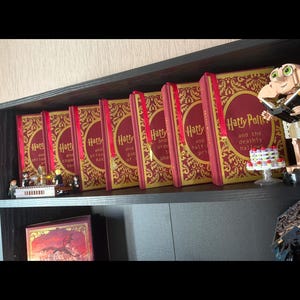 May include: A display of Harry Potter books with red covers and gold lettering, arranged on a shelf. The books are lined up, with decorative items such as a Lego figure and a small cake adding visual interest to the display.
