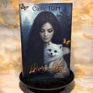 May include: A book cover with a woman and a white fluffy dog. The title "Brimstone" is in gold script, with the author's name, "Callie Hart," above. Two butterfly illustrations are on the cover. The book is displayed on a black stand.