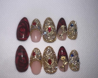 May include: A set of ten artificial nails in various designs. Some nails feature a deep red swirl pattern, while others are adorned with gold embellishments, rhinestones, and a rose design. The nails are oval-shaped.