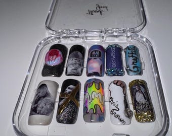May include: A set of ten press-on nails in a clear plastic case. The nails feature various designs, including images, glitter, and text. The color palette includes black, white, gold, and iridescent hues. Some nails have the text "Chris Brown".