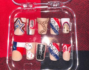 May include: A clear plastic case containing ten false fingernails. The nails are decorated with various designs, including a cowboy hat, a Texas flag, playing cards, and the words "She's Comin'" and "Garite".
