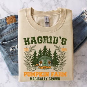 May include: A beige sweatshirt with the text "Hagrid's Pumpkin Farm" in green and orange. The design includes a small house, trees, and pumpkins. The sweatshirt is folded on top of a pair of blue jeans.
