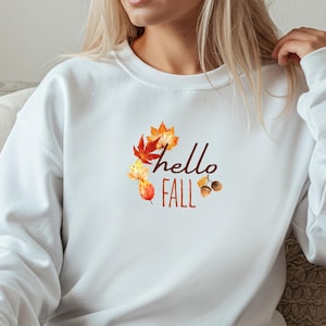 Könnte beinhalten: Ein weißes Sweatshirt mit Rundhalsausschnitt und einem zentralen Grafikdesign mit dem Schriftzug "hello FALL" in brauner Schreib- und Blockschrift, umgeben von einer Ansammlung herbstlicher Blätter im Aquarellstil in Orange, Rot und Gelb sowie zwei Eicheln.