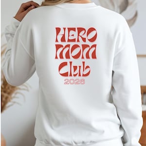 Könnte beinhalten: Weißes Sweatshirt mit dem roten Aufdruck "HERO MOM CLUB 2026" auf dem Rücken. Der Text hat eine Retro-Schriftart mit geschwungenen Buchstaben. Das Sweatshirt ist ein lässiger, bequemer Stil.