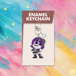以下が含まれることがあります： 紫色の髪と黒い衣装の漫画のキャラクターが特徴の、エナメルキーホルダー。キーホルダーにはシルバーの留め金と、文字「A」の小さなチャームが付いています。パッケージには「ENAMEL KEYCHAIN」と書かれています。