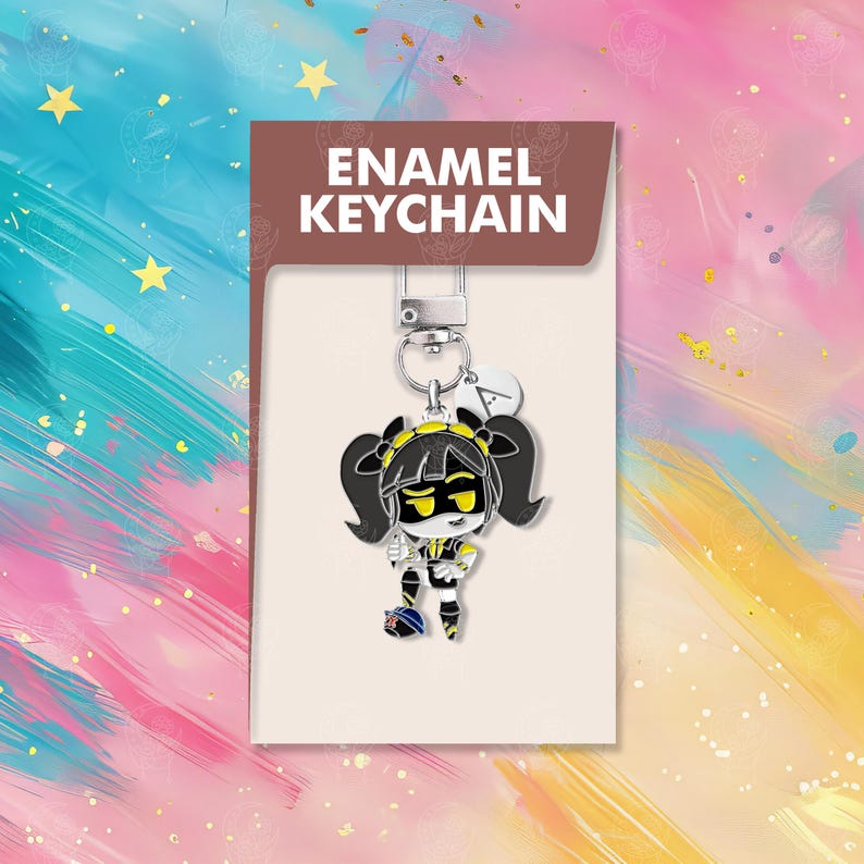以下が含まれることがあります： 黒い髪と黄色のアクセントを持つ漫画のキャラクターが特徴の、エナメルキーホルダー。キーホルダーはシルバーの留め金と小さなチャームに付いています。カードには「ENAMEL KEYCHAIN」と書かれています。