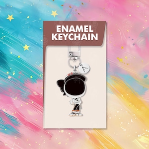 以下が含まれることがあります： オレンジ色のアクセントが付いた白黒の制服を着た漫画のキャラクターが特徴の、エナメルキーホルダー。キーホルダーにはシルバーの留め金と、文字「A」の小さなチャームが付いています。製品カードには、茶色の背景に白い文字で「ENAMEL KEYCHAIN」と書かれています。