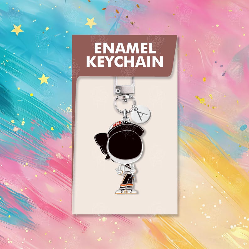 以下が含まれることがあります： オレンジ色のアクセントが付いた白黒の制服を着た漫画のキャラクターが特徴の、エナメルキーホルダー。キーホルダーにはシルバーの留め金と、文字「A」の小さなチャームが付いています。製品カードには、茶色の背景に白い文字で「ENAMEL KEYCHAIN」と書かれています。