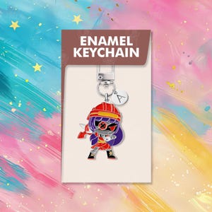 以下が含まれることがあります： 赤、紫、黒の漫画のキャラクターが特徴の、エナメルキーホルダー。キーホルダーはシルバーカラーの留め金と、文字「A」の小さなチャームが付いています。パッケージには「ENAMEL KEYCHAIN」と書かれています。