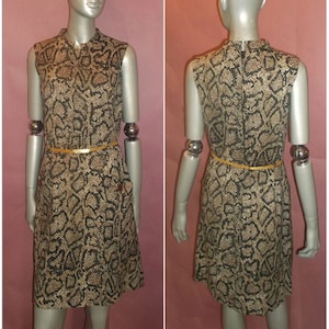 Könnte beinhalten: Ärmelloses Kleid mit Schlangenmuster in Braun-, Beige- und Schwarztönen. Das Kleid hat einen Kragen, ein figurbetontes Oberteil und einen ausgestellten Rock. Ein dünner, goldfarbener Gürtel schnürt die Taille. Das Kleid wird auf einer Schaufensterpuppe präsentiert.