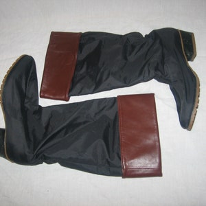May include: A pair of black knee-high boots with brown leather-like cuffs. The boots have a low heel and a rubber sole. The boots are laying on a white surface.