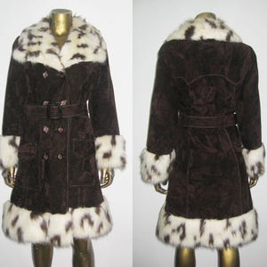 May include: A brown suede coat with a white and brown spotted faux fur collar, cuffs, and hem. The coat features a double-breasted design, a belt at the waist, and two front pockets. The coat is displayed on a mannequin.