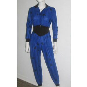 May include: A blue jumpsuit with a black waist belt and black leaf-like patterns. The long-sleeved jumpsuit has a collared neckline and a zipper closure. The cuffs and collar are black. The jumpsuit is displayed on a mannequin.