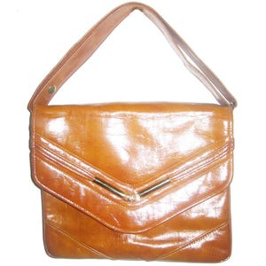 May include: A vintage brown leather handbag with a shoulder strap. The bag has a rectangular shape with a flap closure and a gold-colored metal bar detail. The leather has a glossy finish and visible stitching.