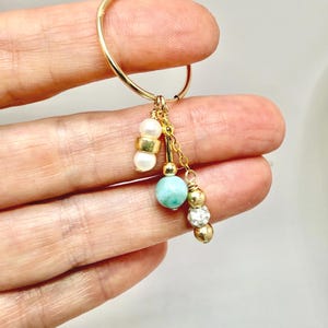 May include: A gold hoop earring featuring three dangling charms. One charm has two white pearls separated by a gold bead. The second is a single light blue stone bead. The third is a gold chain with two gold beads and a clear faceted bead.