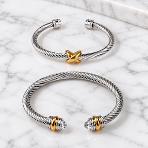 May include: Two open cuff bracelets featuring a twisted cable design in silver-tone metal. The top bracelet has a central gold-tone X-shaped accent. The bottom bracelet features gold-tone end caps with clear, faceted stones at each opening.