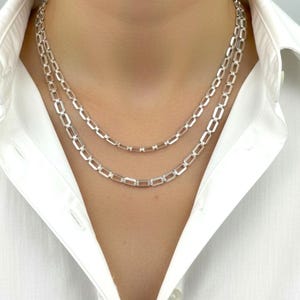 May include: Two silver-colored chain necklaces with rectangular links. The necklaces are layered, with one slightly longer than the other. The chains are displayed against a white collared shirt.