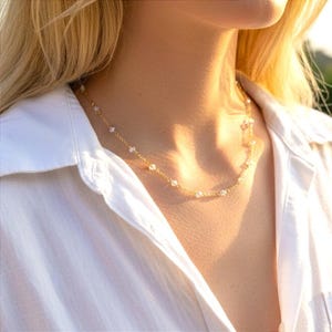 Könnte beinhalten: Eine zarte goldene Kette mit gleichmäßig verteilten kleinen weißen Perlen. Die Halskette liegt am Hals an und zeigt ein feines Metallkettendesign mit reflektierenden, kreisförmigen Akzenten, die über die gesamte Länge der Kette verteilt sind.