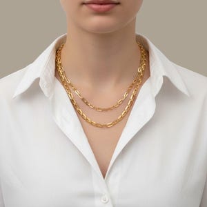 May include: Two gold-colored chain necklaces with rectangular links are worn over a white collared shirt. The necklaces are different lengths, creating a layered effect. The background is a neutral, light gray.