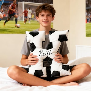 May include: A black and white football-shaped cushion in the form of the letter "K", with the name "Keith" printed on it. The cushion is held by a person sitting on a bed. Football-themed decor is visible in the background.
