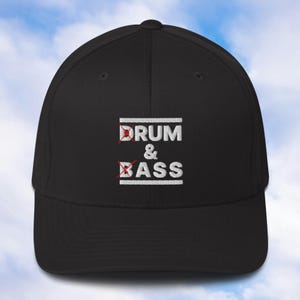 以下が含まれることがあります： 黒い野球帽。白い文字で「DRUM & BASS」と刺繍されています。テキストは白い線で囲まれ、「DRUM」と「BASS」の上に赤いバツ印があります。帽子はカーブしたつばと構造的なフィット感を持っています。